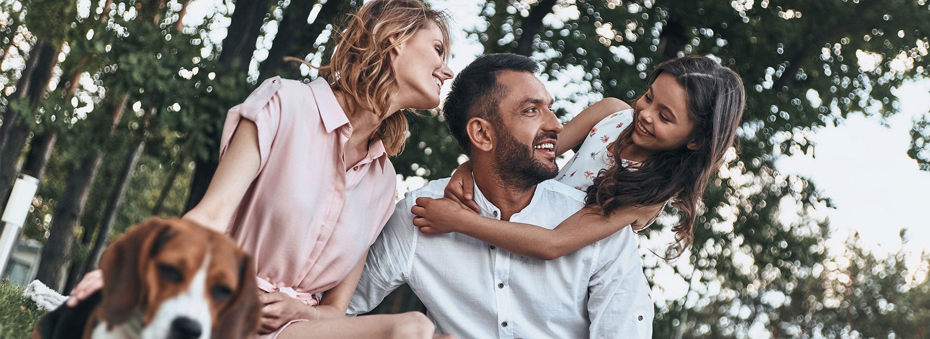 A family sitting together outdoors in a leafy park — a woman and a man smiling as a young girl hugs the man from behind, with a beagle dog resting beside them.